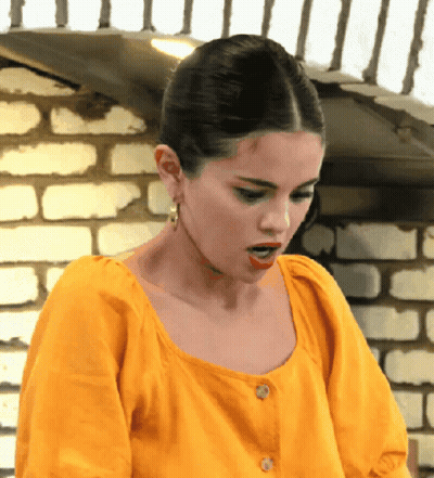 A Woman In A Yellow Shirt Is Standing In Front Of A Brick Wall With Her Mouth Open GIF