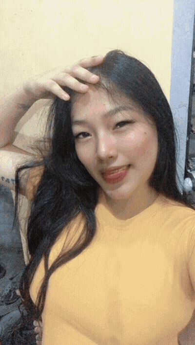 A Woman In A Yellow Shirt Is Taking A Selfie With Her Hand On Her Forehead GIF