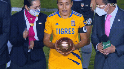 A Woman In A Yellow Shirt That Says Tigres Is Holding A Gold Ball GIF