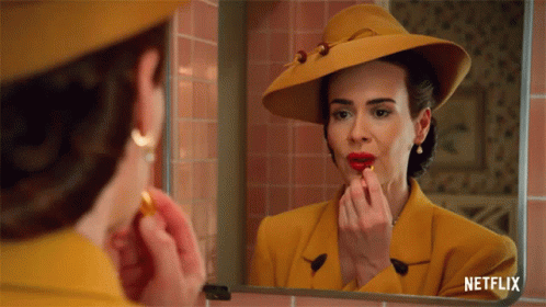 A Woman In A Yellow Suit And Hat Is Applying Red Lipstick GIF