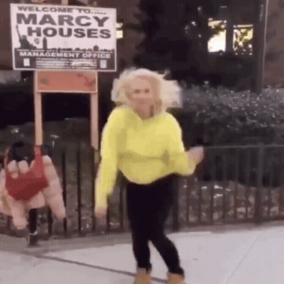 A Woman In A Yellow Sweater Is Dancing In Front Of A Marcy Houses Sign . GIF