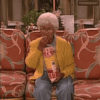 A Woman In A Yellow Sweater Is Sitting On A Couch Holding A Bag Of Popcorn GIF