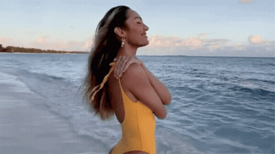 A Woman In A Yellow Swimsuit Is Standing On The Beach Looking At The Ocean . GIF