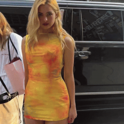 A Woman In A Yellow Tie Dye Dress Stands In Front Of A Black Van That Says Dsquared On The Side GIF
