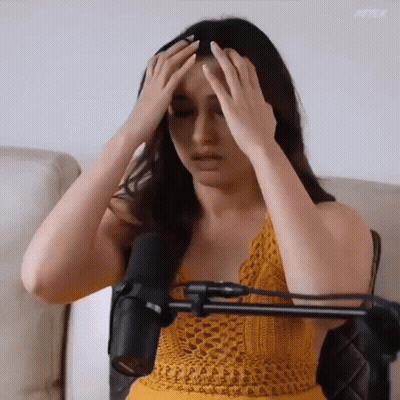 A Woman In A Yellow Top Is Sitting In Front Of A Microphone With Her Hands On Her Head GIF