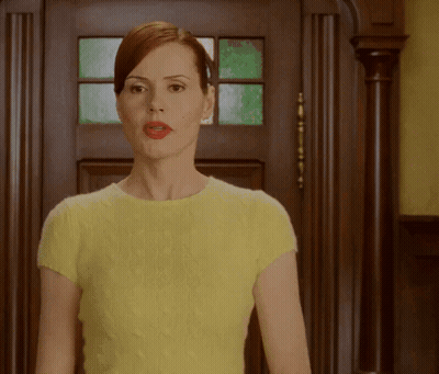 A Woman In A Yellow Top Is Standing In Front Of A Wooden Door GIF