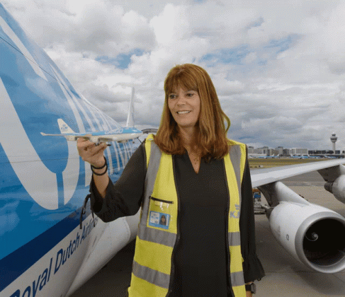 A Woman In A Yellow Vest Is Standing In Front Of A Royal Dutch Airlines Plane GIF