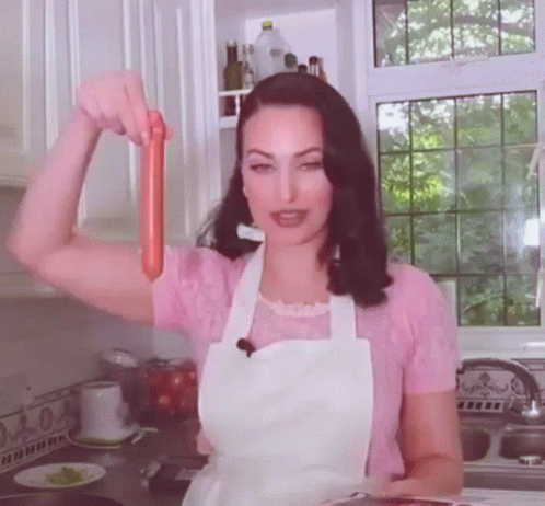 A Woman In An Apron Holds A Sausage In Her Hand GIF