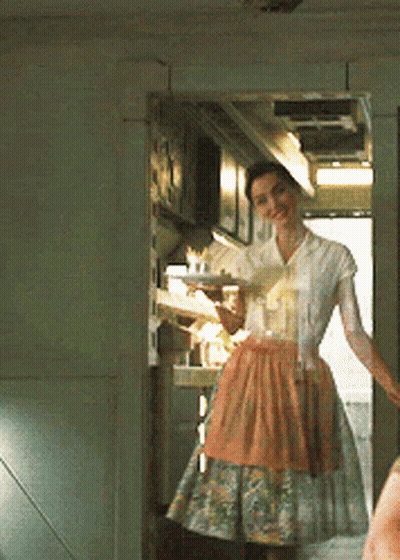 A Woman In An Apron Is Standing In A Kitchen Holding A Tray GIF