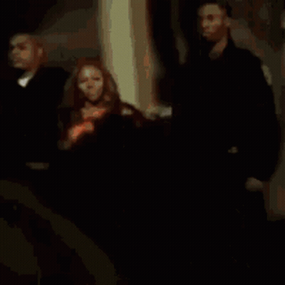 A Woman In An Orange Dress Is Standing Between Two Men In Black Suits GIF