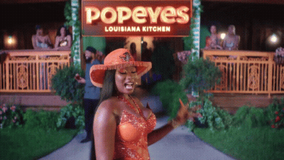 A Woman In An Orange Dress Is Standing In Front Of A Popeye 'S Louisiana Kitchen Sign GIF