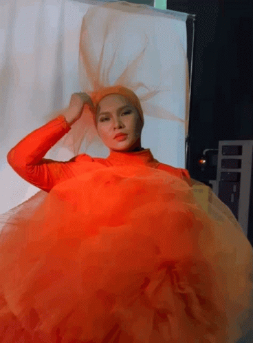 A Woman In An Orange Dress Is Standing In Front Of A White Backdrop GIF