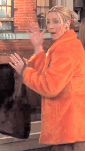 A Woman In An Orange Fur Coat Is Standing In Front Of A Brick Building And Waving . GIF