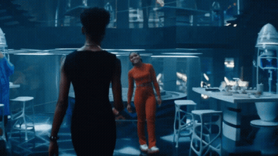 A Woman In An Orange Jumpsuit Stands Next To Another Woman In A Black Dress GIF