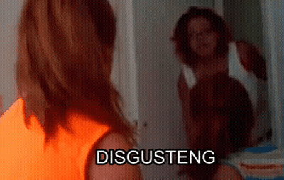 A Woman In An Orange Shirt Is Standing In Front Of A Mirror With The Word Disgusteng On It GIF