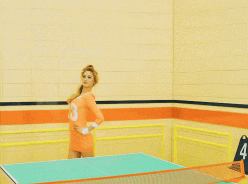 A Woman In An Orange Shirt With The Number 8 On It Is Standing On A Ping Pong Table GIF