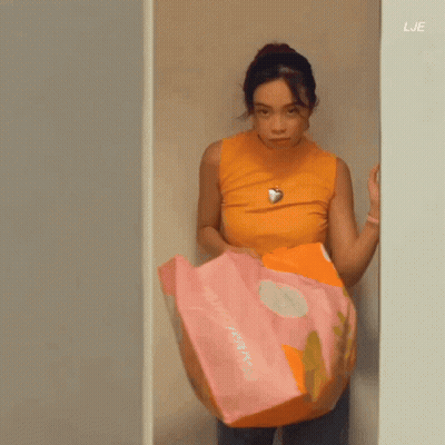 A Woman In An Orange Tank Top Is Standing In A Closet Holding A Pink And Orange Blanket . GIF