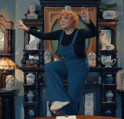 A Woman In Blue Overalls Stands On One Leg In Front Of A Fireplace GIF