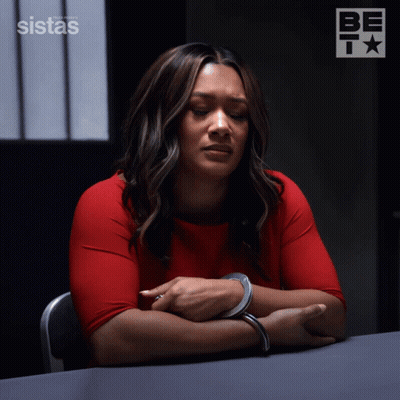 A Woman In Handcuffs Sits At A Table In Front Of A Sign That Says Sistas On It GIF
