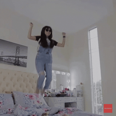 A Woman In Overalls Is Jumping In The Air In A Bedroom With A Picture Of A Bridge On The Wall GIF