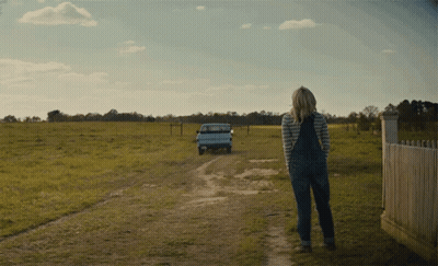 A Woman In Overalls Is Walking Down A Dirt Road With A Truck Behind Her GIF