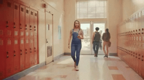 A Woman In Overalls Is Walking Down A Hallway In A School . GIF