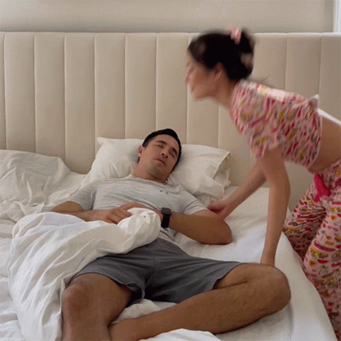 A Woman In Pink Pajamas Is Standing Next To A Man In A Bed GIF