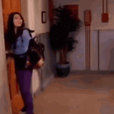 A Woman In Purple Pants Is Standing In A Hallway Holding A Backpack . GIF