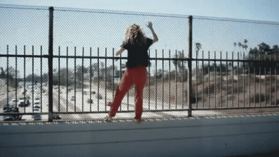 A Woman In Red Pants Is Standing On A Bridge Looking Over A Fence GIF