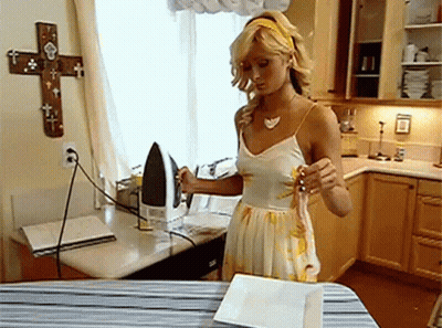 A Woman Ironing Clothes In A Kitchen With A Cross On The Wall Behind Her GIF