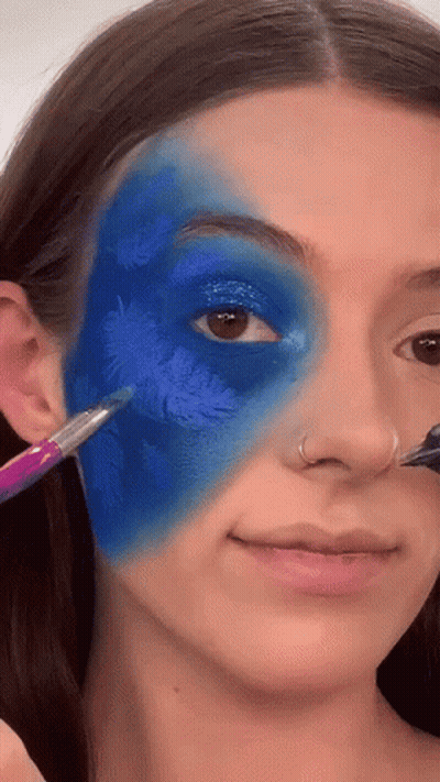 A Woman Is Applying Blue Paint To Her Face With A Brush . GIF