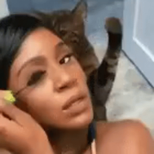 A Woman Is Applying Mascara To Her Eyelashes While A Cat Sits On Her Head . GIF