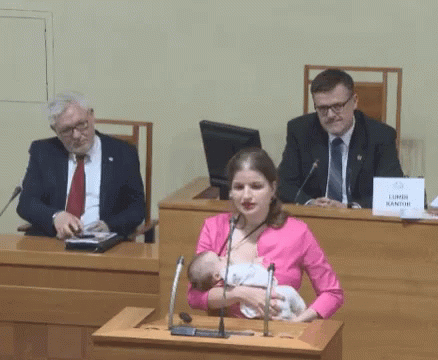 A Woman Is Breastfeeding A Baby In Front Of A Podium With A Sign That Says Lumen Kantor GIF