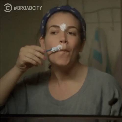 A Woman Is Brushing Her Teeth In Front Of A Mirror With Broad City Written On The Bottom GIF