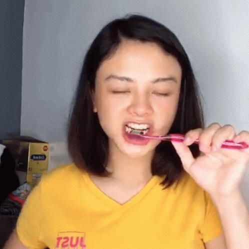 A Woman Is Brushing Her Teeth With A Pink Toothbrush And Wearing A Yellow Shirt That Says T2ul GIF