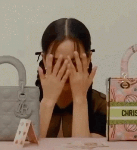 A Woman Is Covering Her Face With Her Hands While Sitting At A Table With Handbags . GIF