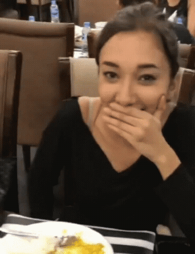 A Woman Is Covering Her Mouth With Her Hand While Sitting At A Table GIF