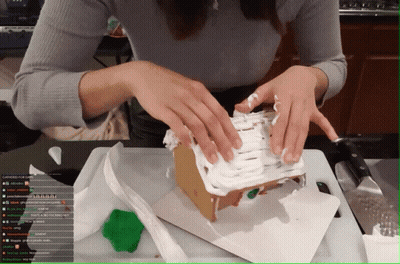 A Woman Is Decorating A Gingerbread House On A Cutting Board While Watching A Stream On Youtube GIF