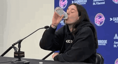 A Woman Is Drinking Water From A Bottle While Sitting At A Table With A Microphone . GIF