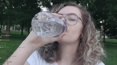 A Woman Is Drinking Water From A Plastic Bottle In Front Of Trees GIF