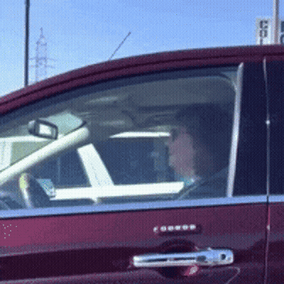 A Woman Is Driving A Red Car With A Sign In The Background That Says ' Coca Cola ' GIF