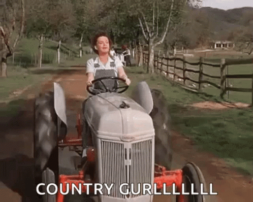 A Woman Is Driving A Tractor Down A Dirt Road In A Field . GIF