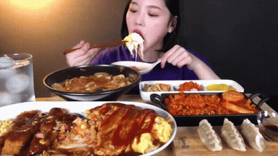 A Woman Is Eating A Plate Of Food With A Spoon And Chopsticks While Sitting At A Table GIF