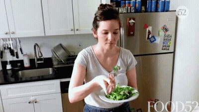 A Woman Is Eating A Salad In A Kitchen With A Sign That Says What It Is On The Refrigerator GIF