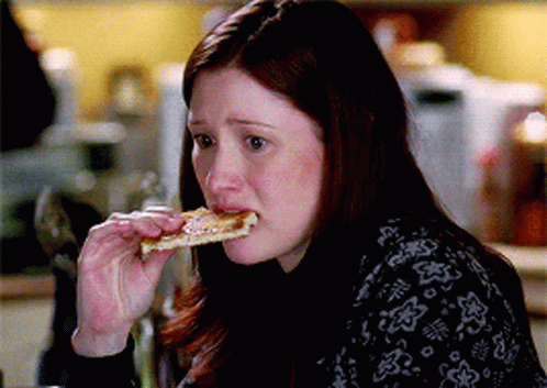 A Woman Is Eating A Slice Of Pizza While Wearing A Black Sweater With Flowers On It GIF