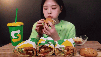 A Woman Is Eating A Subway Sandwich Next To A Cup That Says ' S ' On It GIF