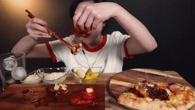 A Woman Is Eating Food With Chopsticks And A Pizza On A Wooden Cutting Board GIF