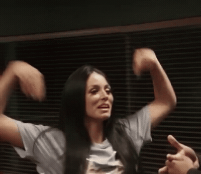 A Woman Is Flexing Her Muscles In Front Of A Window While Wearing A White Shirt . GIF