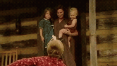 A Woman Is Holding A Baby And A Dog In Her Arms While Standing Next To A Log Cabin . GIF