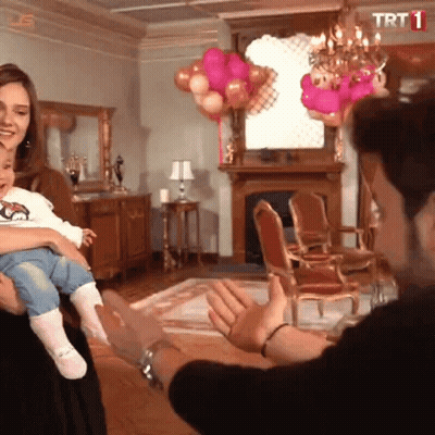 A Woman Is Holding A Baby In A Living Room With Balloons And A Trt Logo GIF